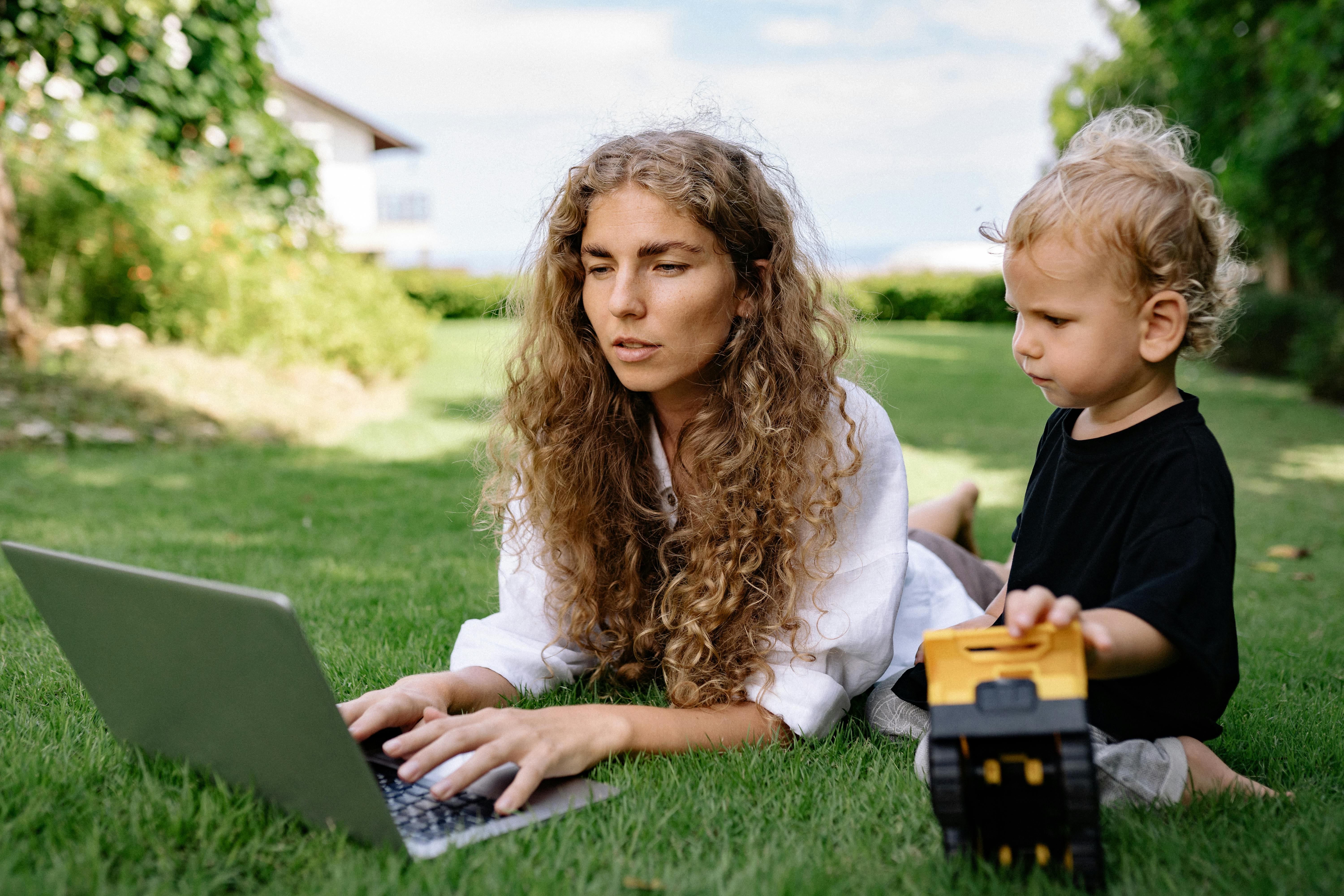 Woman working on a laptop while lying on grass with her child nearby playing