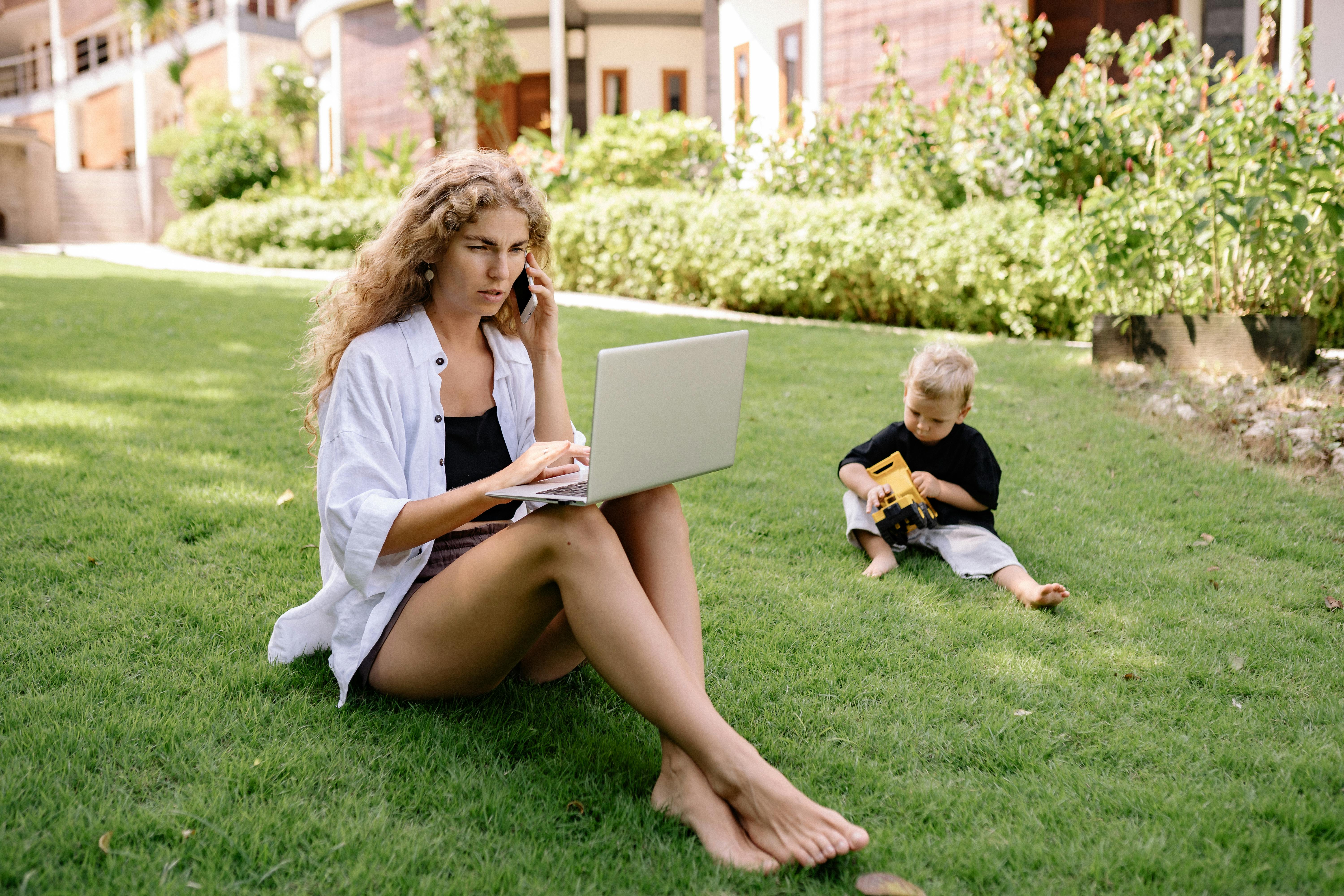 photo of woman using laptop while sitting on grass field with her child