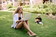 一名妇女在户外用笔记本电脑远程工作，她的孩子在附近的草地上玩耍