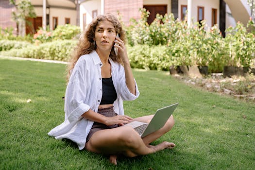 A woman in casual attire works remotely on a laptop and phone while sitting on grass.