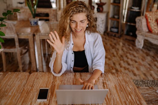 Smiling woman waves during a video call on her laptop in a comfortable indoor setting.