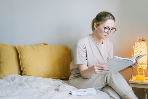 Free Young woman reading a book while sitting on a bed in a cozy, relaxed bedroom environment. Stock Photo
