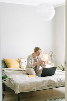 A focused woman works on her laptop while sitting on her bed in a cozy bedroom.