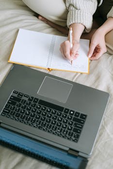 Close-up image of hands writing in a notebook beside a laptop, capturing a cozy learning environment.
