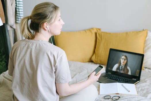 Woman sitting on bed in a home setting, working on laptop with smartphone and headphones.