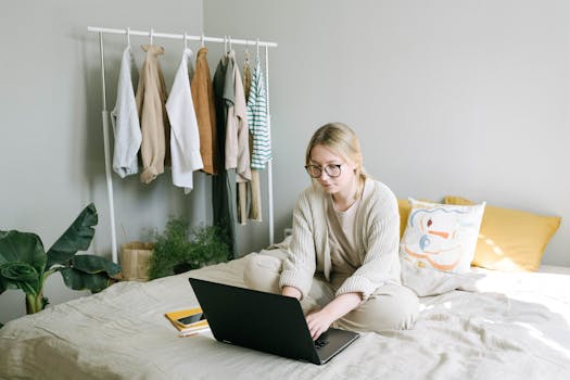 Caucasian woman with eyeglasses working on laptop in comfortable bedroom setting, surrounded by clothes and plants.
