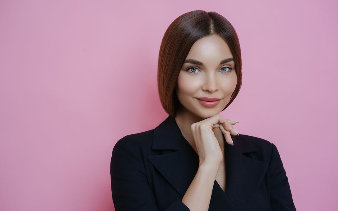 Happy female near pink wall and smiling while looking at camera and touching chin