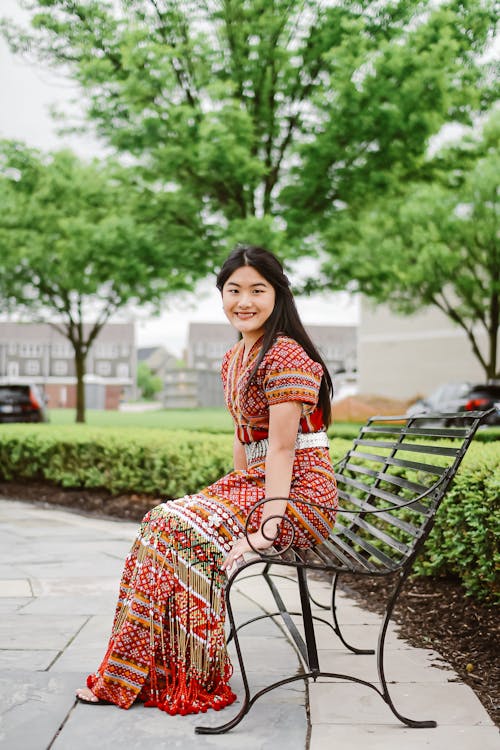 Optimistic Asian female in dress sitting on bench in park and looking at camera with toothy smile