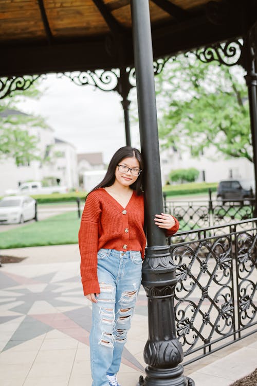 Free Positive young Asian female in casual outfit and eyeglasses standing in ornamental roofed terrace in sunny park Stock Photo