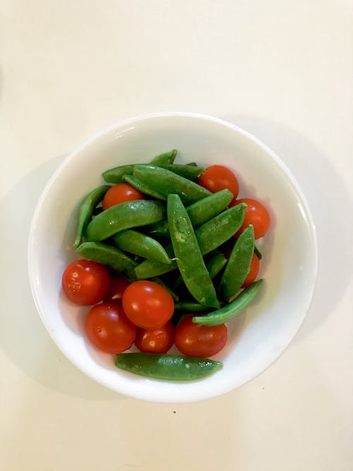 Top view of fresh ripe cherry tomatoes placed in white ceramic bowl with organic unpeeled green peas on white table