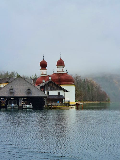 Aged Catholic St. Bartholomew Church with brown tiled cupolas located on calm pond shore on foggy day in Germany