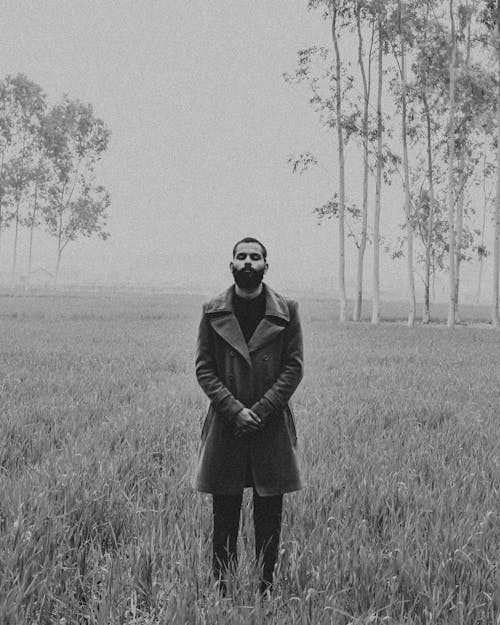 Black and white contemplative ethnic male in stylish coat standing with hands folded and eyes closed foggy nature
