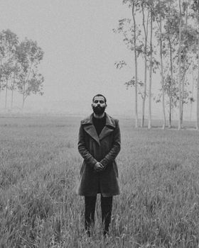 Black and white contemplative ethnic male in stylish coat standing with hands folded and eyes closed foggy nature