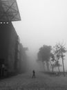 Black and white distant person walking on pavement near big industrial building on foggy gloomy day