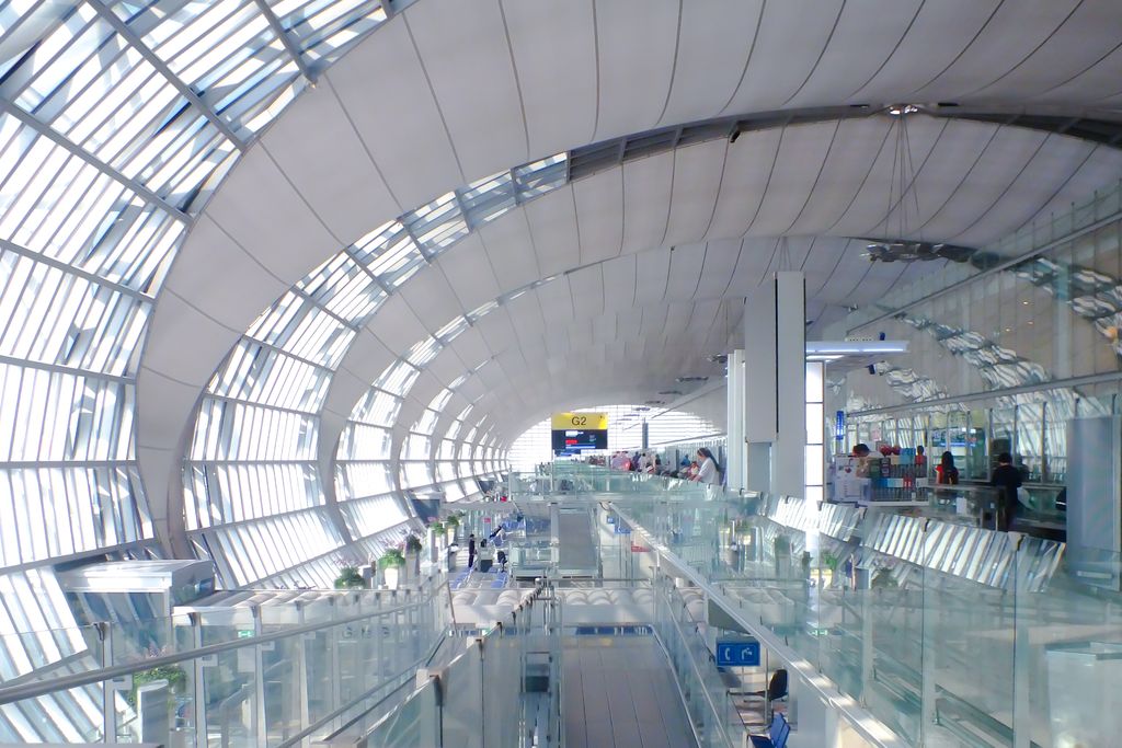 An airport terminal concourse under a long barrel-vaulted roof of white ribs and glass, with glazed balustrades down both sides and travellers in the distance.