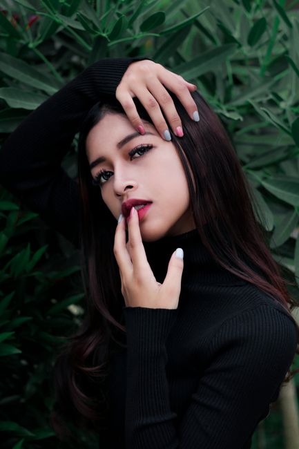 Charming young lady in black turtleneck sweater touching gently lips and face while standing against lush green foliage and looking at camera