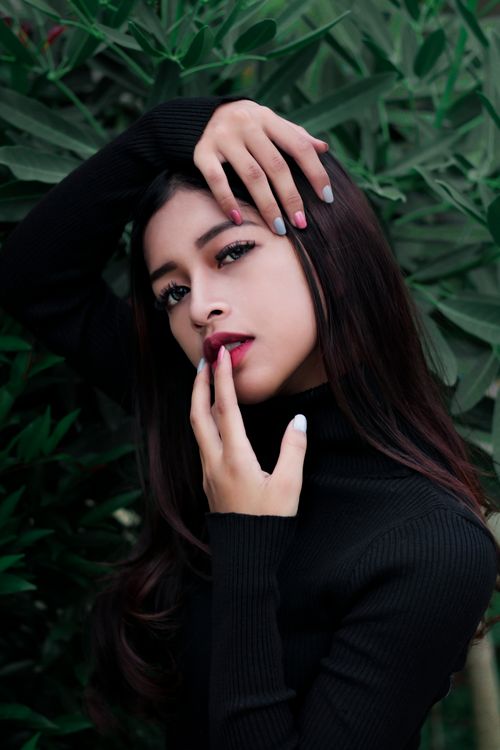 Free Charming young lady in black turtleneck sweater touching gently lips and face while standing against lush green foliage and looking at camera Stock Photo