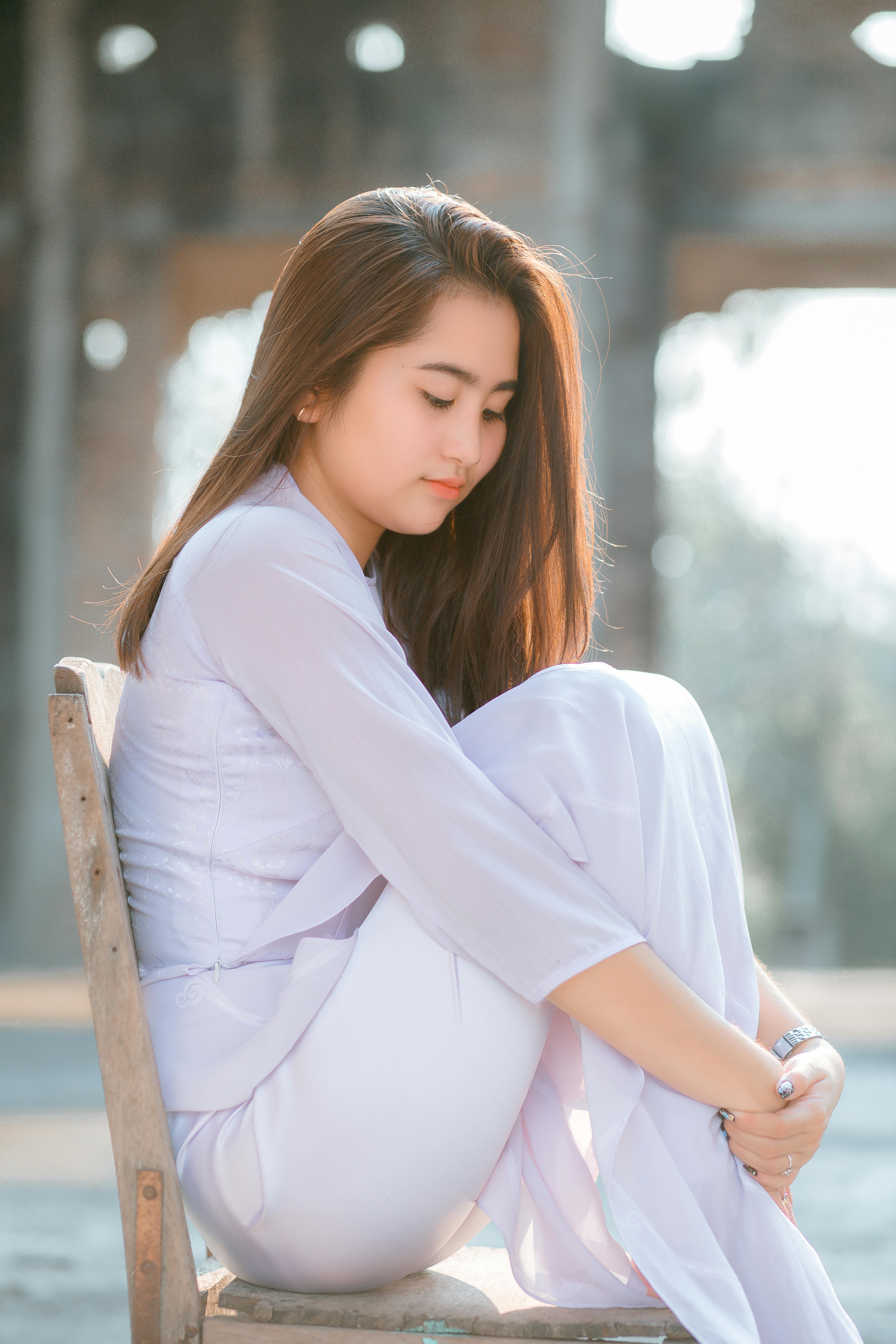 Side view full length charming young Asian lady in white clothes sitting on chair on sunny street and hugging knees while looking down modestly