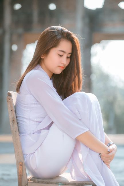 Side view full length charming young Asian lady in white clothes sitting on chair on sunny street and hugging knees while looking down modestly