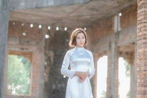 Free Elegant young Asian female wearing silk white dress standing in remaining brick building and looking away dreamily Stock Photo