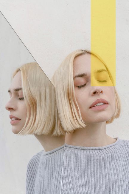Stylish young female with blond hair in casual sweater standing near mirror with closed eyes