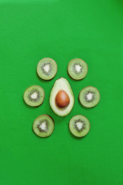 Top view of halved fresh avocado placed on green table with slices of ripe kiwi