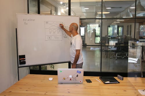 Side view of anonymous hairless male employee in white t shirt drawing draft on whiteboard while working on startup strategy in contemporary office