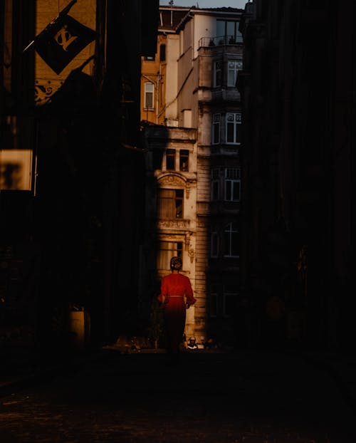 Back view of unrecognizable female in red dress walking along narrow street in old residential district at sunset