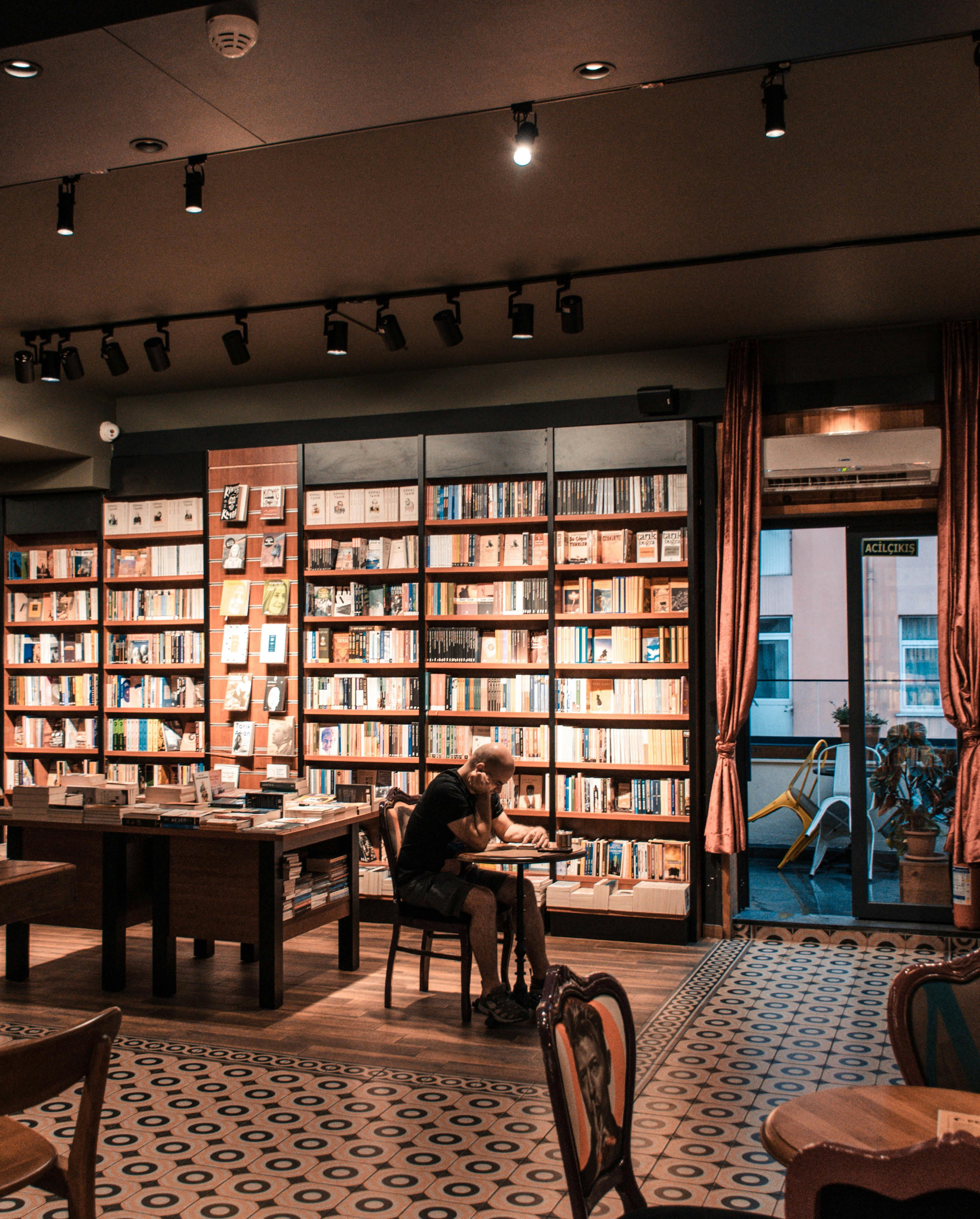 Free A man sits reading in a cozy bookstore, surrounded by shelves filled with books, capturing a warm and intellectual atmosphere. Stock Photo
