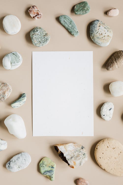 Free Top view composition of assorted whole and cracked pebbles arranged around white empty paper sheet on beige surface Stock Photo