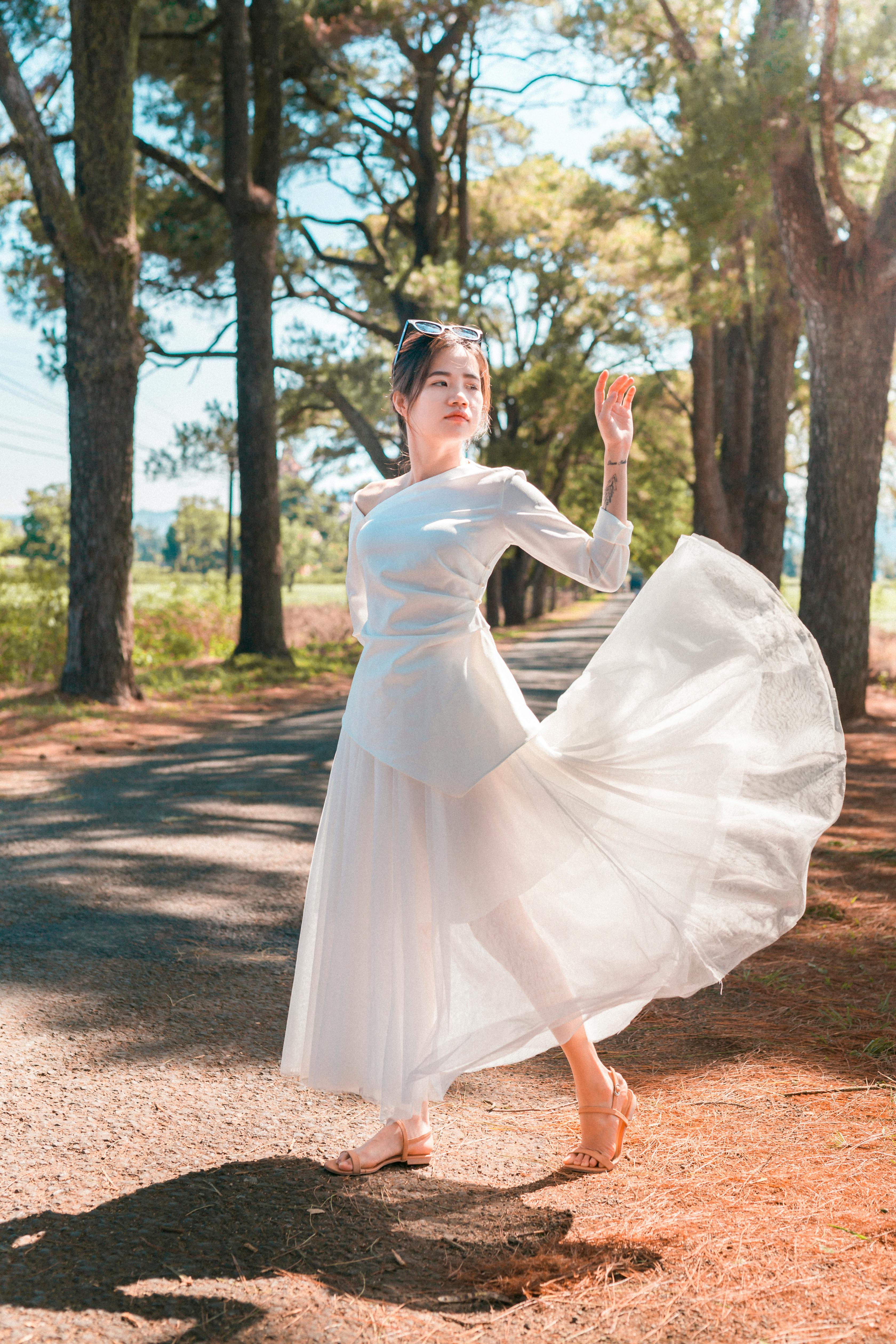 阳光明媚的日子里，一位身着白色连衣裙的女子站在公园的树林中，举着手