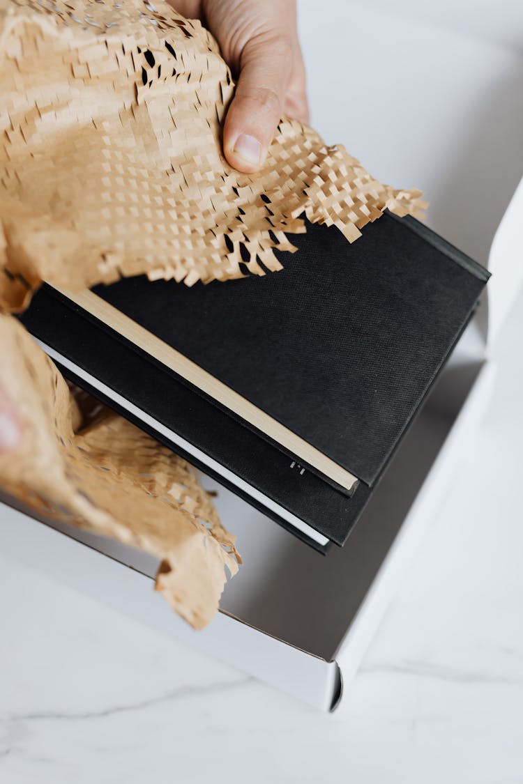 Crop Man Wrapping Stack Of Black Notebooks In Packing Paper