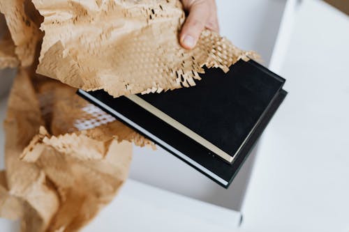 From above unrecognizable adult male taking black notebooks with craft perforated paper under white carton box while preparing mail delivery at white table