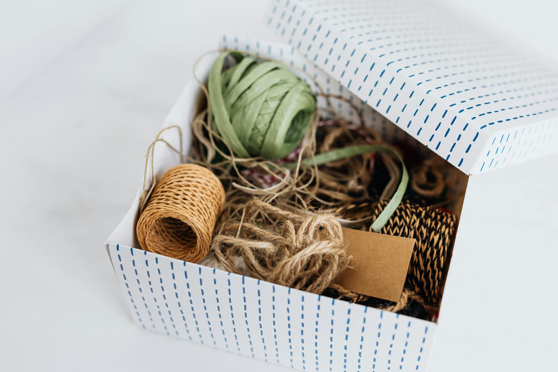 From above open carton box filled with various ribbons and ropes for holiday presents and gifts wrapping and blank brown cardboard card placed on white table