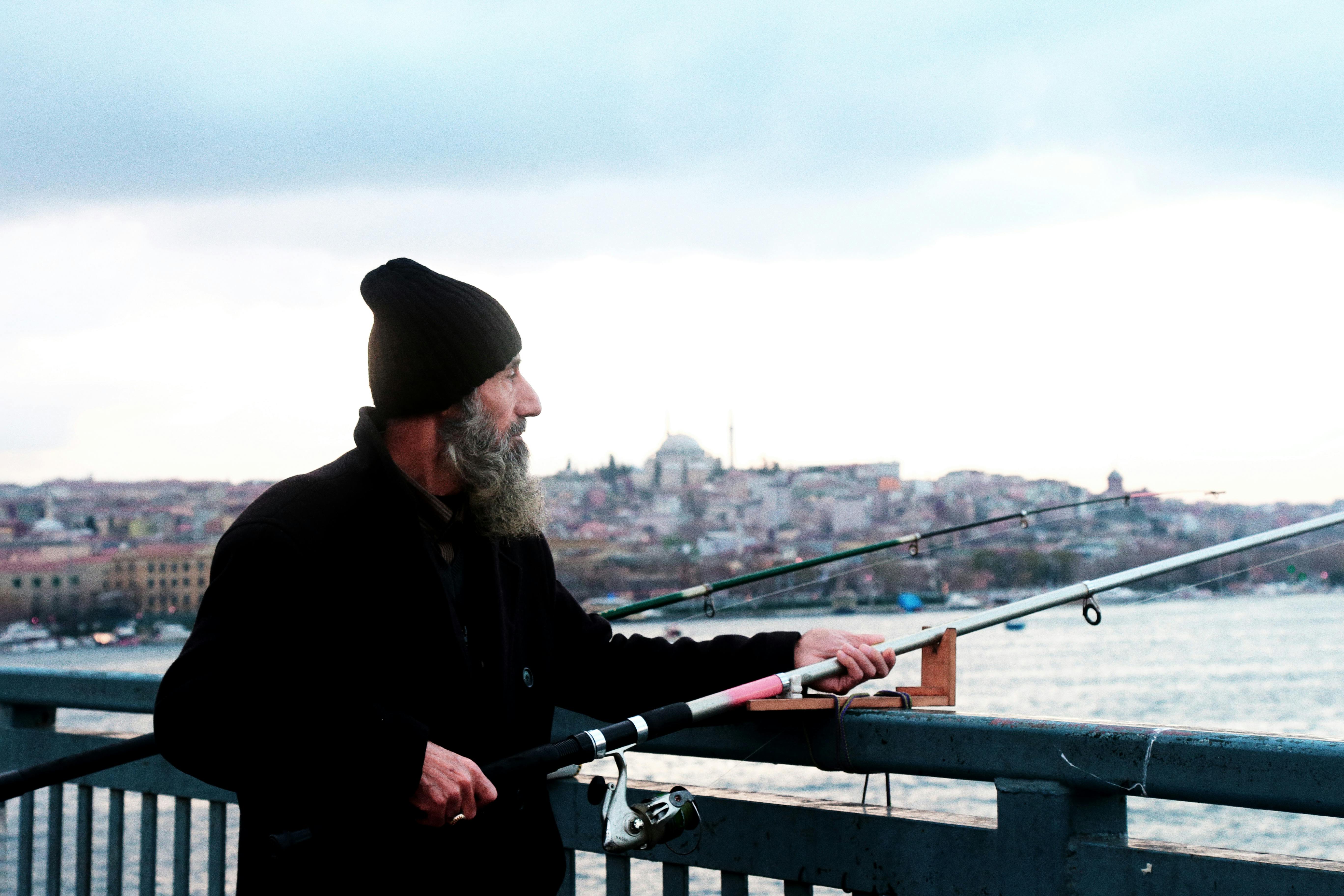 Bearded elderly man fishing from a bridge in Istanbul, Turkey with a cityscape backdrop.