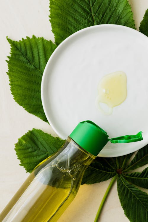 Free Top view of open bottle of cosmetic oil placed flat on table near white plate with small amount of oil on it on big fresh green leaf Stock Photo