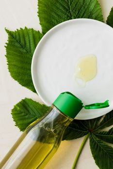 桌子上平放着一个打开的化妆品油瓶，从顶视图看，瓶口靠近白色盘子，盘子上有少量油，整体置于大片新鲜绿叶之上