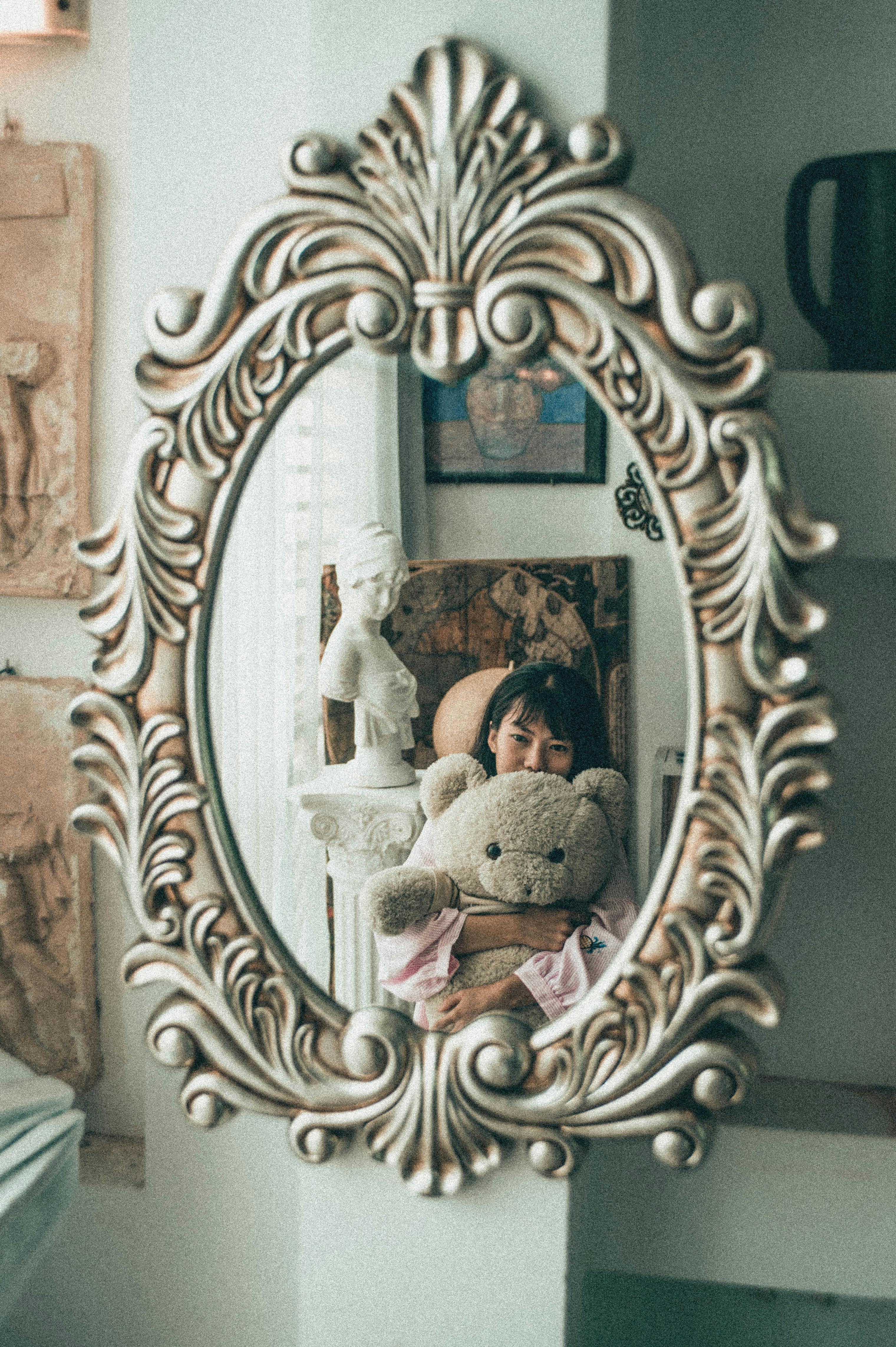 Mirror in elegant massive frame reflecting charming young Asian female cuddling with fluffy bear toy in light bedroom