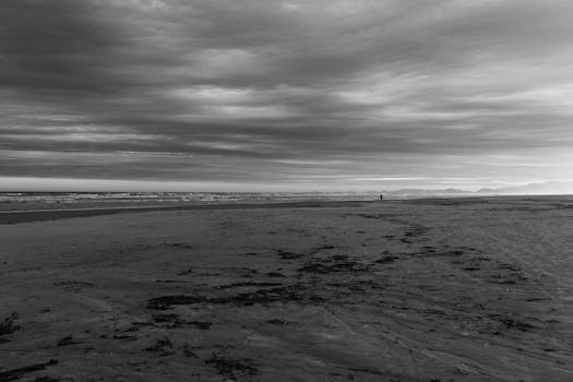 A lone figure on a desolate beach under a dramatic, overcast sky in monochrome.
