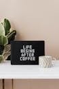 Light up letter message board reading Life Begins After Coffee and ceramic coffee cup on table with green pot plant near pastel pink wall