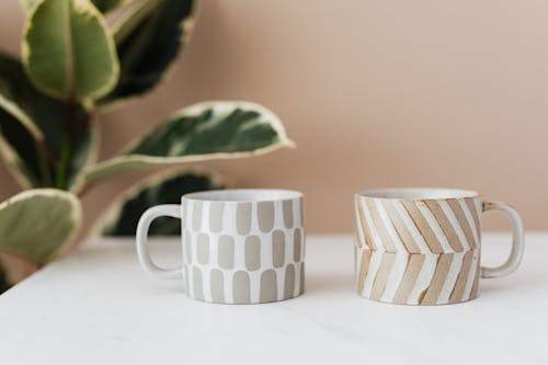 Free Handmade ceramic mugs with creative designs placed on white marble table with blurred green house plant near pink wall in background Stock Photo