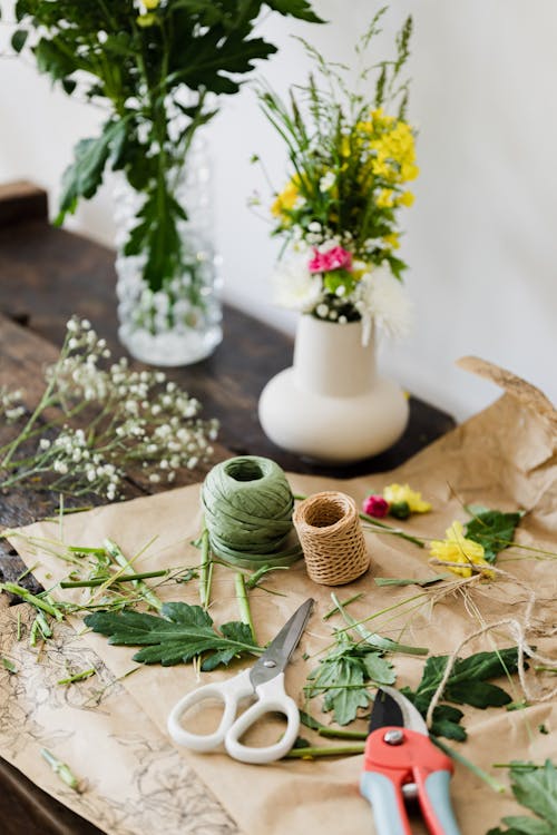 Scissors and pruner together with twine rolls and cut flower leaves near vase bouquets on wooden table at florist workshop