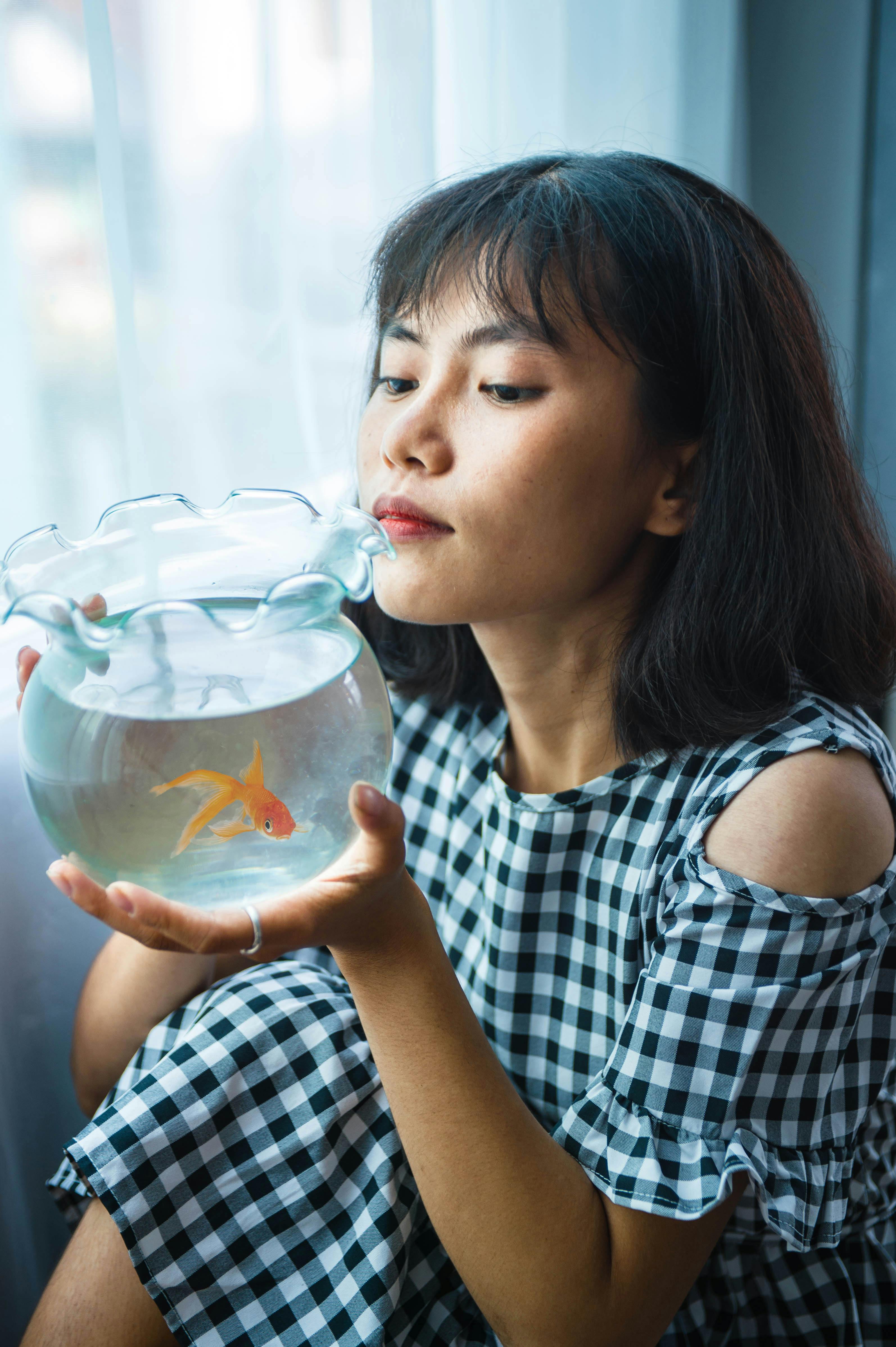 Young Asian female in checkered dress watching tiny goldfish swimming in glass vase while resting near window at home