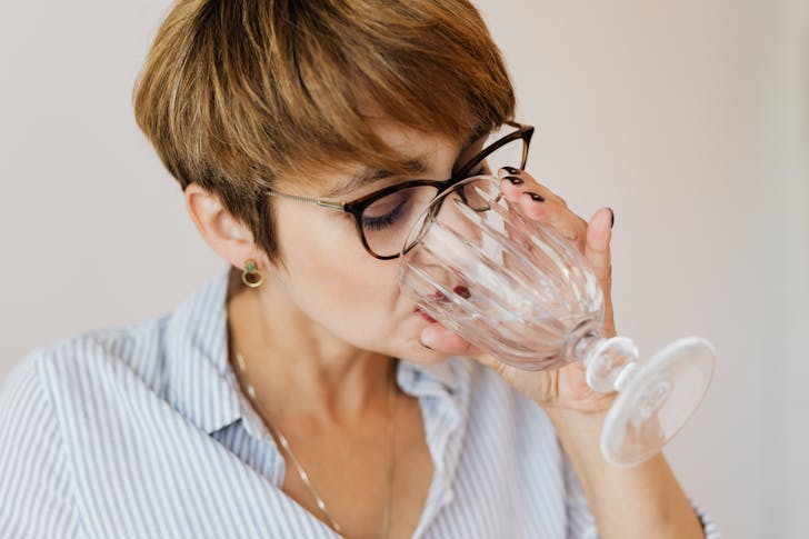 Crop serious female with short hair in shirt with stripes and glasses drinking water while sitting in creative workplace