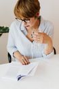 Crop concentrated short haired female in eyeglasses striped shirt sitting at white table near greenery at creative place and reading book