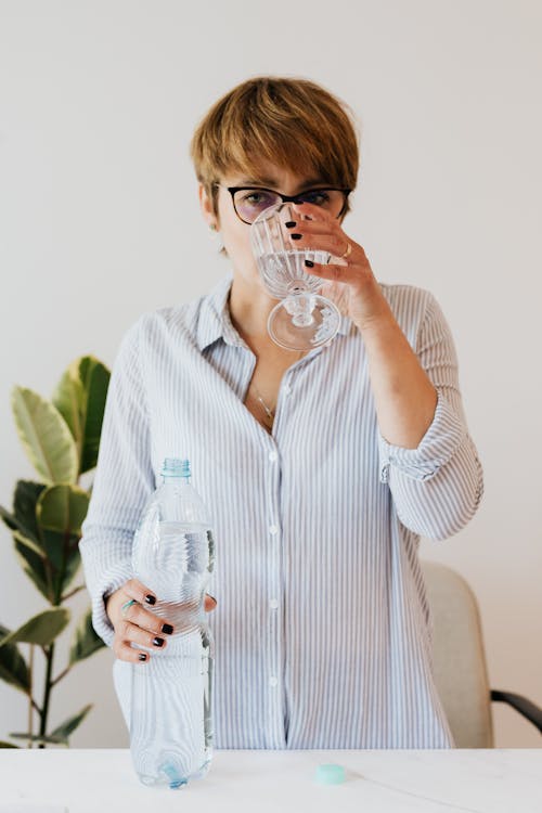 Female with short hair in eyewear and shirt with stripes standing near white table and drinking while holding bottle of water