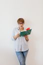 Serious woman with short hair in glasses and jeans with shirt in stripes standing near white wall and reading book