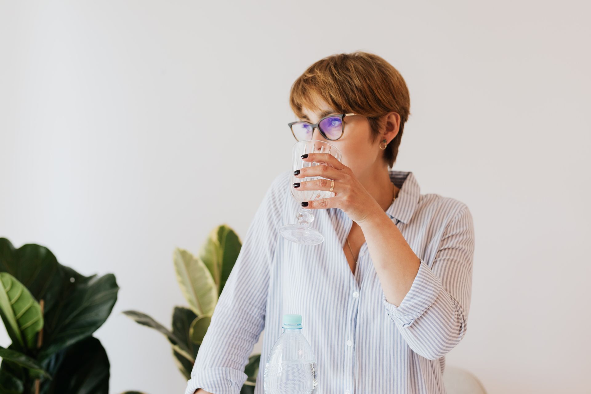 Focused female in casual shirt and eyeglasses drinking pure water from glass while standing against white wall and green houseplants