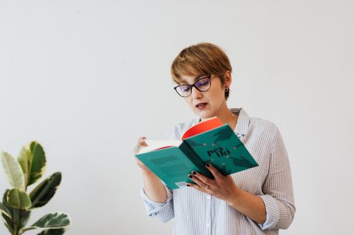 Free Concentrated female in casual shirt and eyeglasses reading interesting book with green hardcover while standing against white wall and houseplant Stock Photo