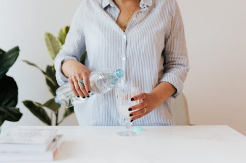 Crop unrecognizable female in casual light blue shirt standing near white table with stack books and pouring pure mineral water from plastic bottle into glass against white wall and plants